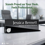 A black glass desk nameplate with white text reading 'Jessica Bennett' and 'Director of Human Resources,' placed on a white office desk with blurred office chairs, a laptop, and a plant in the background. Green text at the top says, 'Stands Proud on Your Desk.