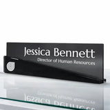 A black acrylic desk nameplate with white text reading 'Jessica Bennett' and 'Director of Human Resources', shown on a reflective white surface.