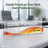 A clear acrylic desk nameplate with a wavy orange and gold gradient base, personalized with the name 'Daniel Thompson' and title 'Senior Project Manager,' shown on a white office desk with blurred office chairs and a window in the background. Large text at the top reads, 'Stands Proud on Your Desk.