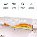 Personalized acrylic business nameplate shown on a light wood surface with dimension callouts and feature icons above. The clear rectangular plaque has a white name, gray subtitle text, and an orange-to-yellow wave design with a black script signature.