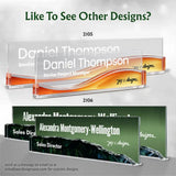 Custom acrylic business nameplates shown in two color/design options: a clear and orange option labeled 2105 and a dark green and black option labeled 2106. The nameplates feature placeholder text for a senior project manager and sales director, with the brand name 'Jay C Designs' visible on the front.