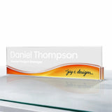 A personalized clear acrylic desktop nameplate sign for 'Daniel Thompson' with a white, orange, and yellow wavy design, standing on a reflective glass surface. Wave like name plate