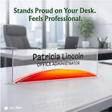 A clear acrylic desk nameplate with a curved red, orange, and yellow gradient base, personalized with the name 'Patricia Lincoln' and the title 'OFFICE ADMINISTRATOR'. The nameplate is displayed on a white desk in a blurred modern office setting with the text 'Stands Proud on Your Desk.