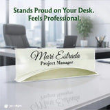 A clear acrylic desk name sign with black printed text reading 'Mari Estrada' and 'Project Manager,' placed on a white office desk with a blurred office background and promotional text above.