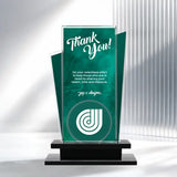 A clear acrylic award plaque with a green printed front panel reading 'Thank You!' and a custom message, displayed on a black tiered pedestal against a white slatted background.