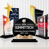 A set of modern acrylic and glass award trophies displayed on black bases against a white paneled background. The central and most prominent trophy is a black semi-circular plaque with gold accents that reads '10TH SUMMIT TECH Celebrating a Decade of Success 2010 - 2025'.