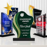 A custom-designed green and gold acrylic award plaque in the center, flanked by other corporate award trophies on a reflective white surface.