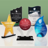 Five acrylic glass award plaques on black bases, including a gold star-shaped Loyalty Award, a red round plaque, a blue teardrop-shaped Employee Recognition Award, and two black rectangular recognition plaques, displayed on a reflective white surface against a green gradient background. Milestone Trophy is centered in the group
