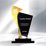 Front view of a modern acrylic and glass award trophy shaped like a stylized abstract figure, with a black front panel reading 'Loyalty Award' and a gold side section with a white circular logo. The trophy sits on a black rectangular base against a white studio background with soft vertical shadows.