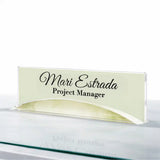 A personalized acrylic nameplate sign with the text 'Mari Estrada Project Manager' in black script and serif font, displayed on a reflective glass surface against a white background.